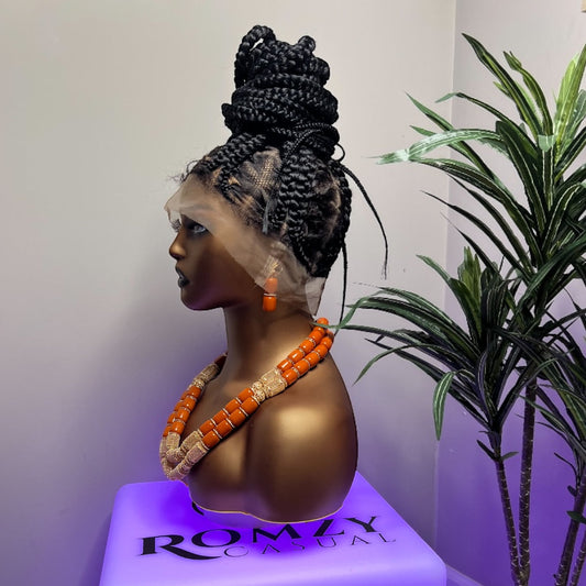 Full lace braided wig on mannequin with top bun, showcasing natural black braids and orange bead necklace next to green plant